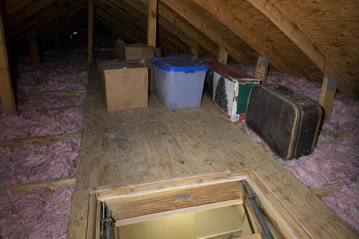 Cardboard boxes and a plastic tote on a plywood walkway in an attic, pink insulation between the joists