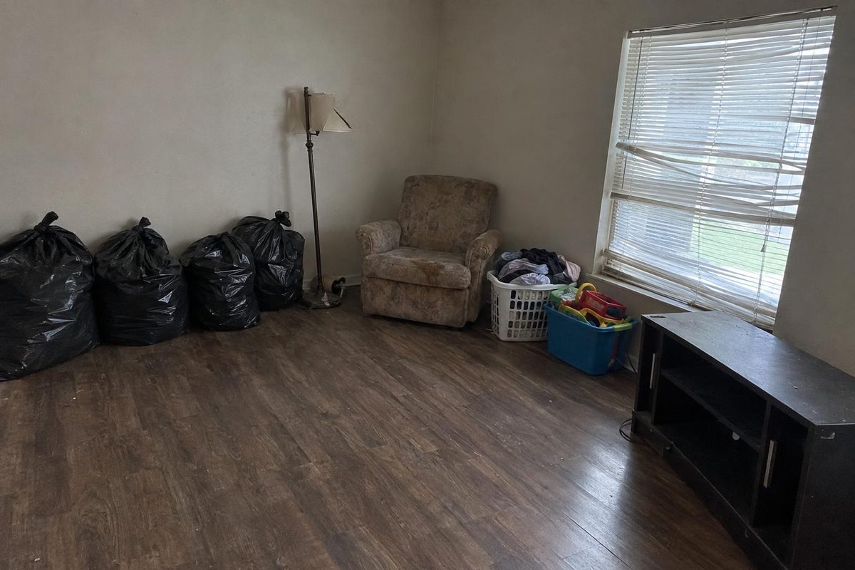 Black trash bags and loose household items lined along the wall of an empty living room