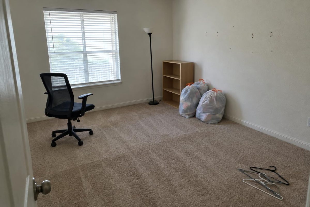 An empty carpeted bedroom with a lamp, a desk chair and a few bags left in the corner