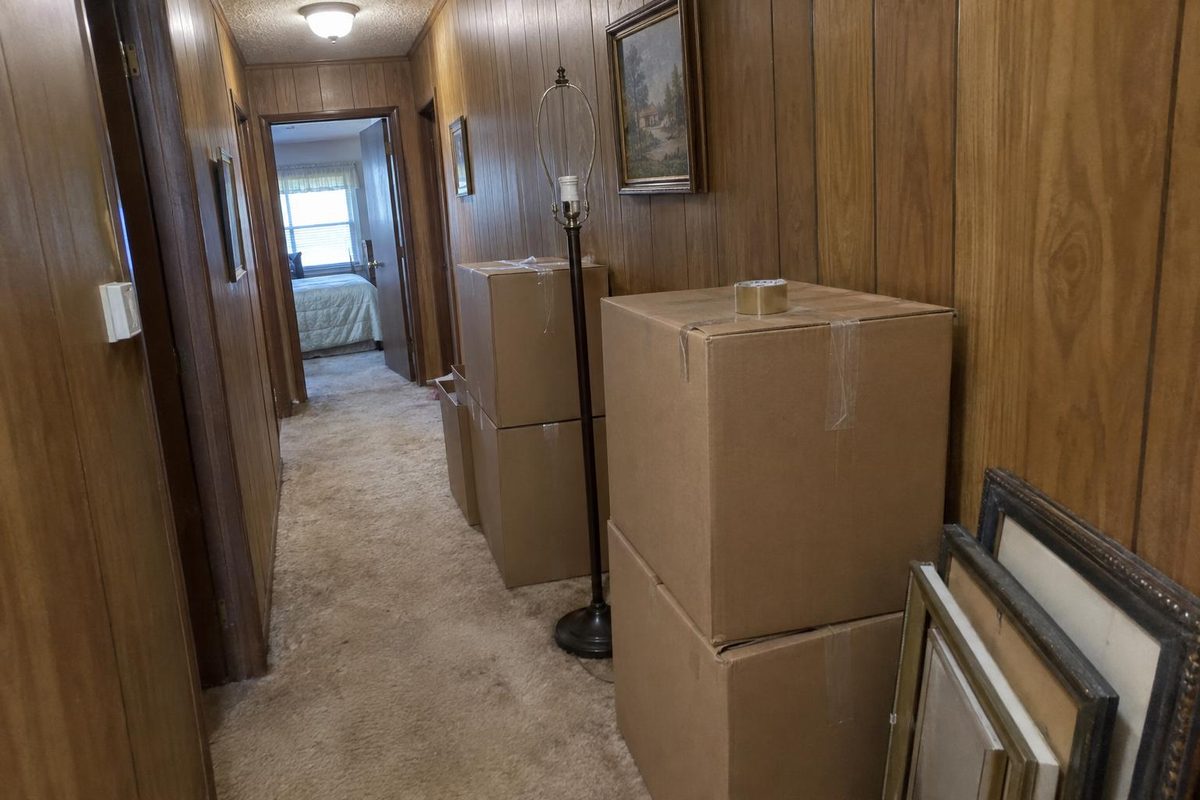 A narrow hallway lined with taped moving boxes and a stack of framed pictures leaning on the wall
