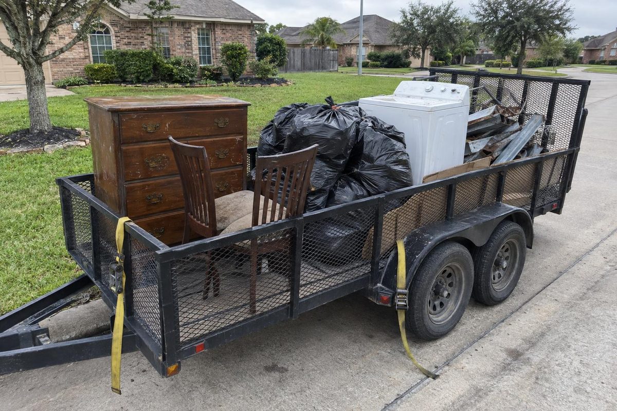 An open utility trailer loaded in sections: furniture at the front, bagged trash in the middle, a washer at the back